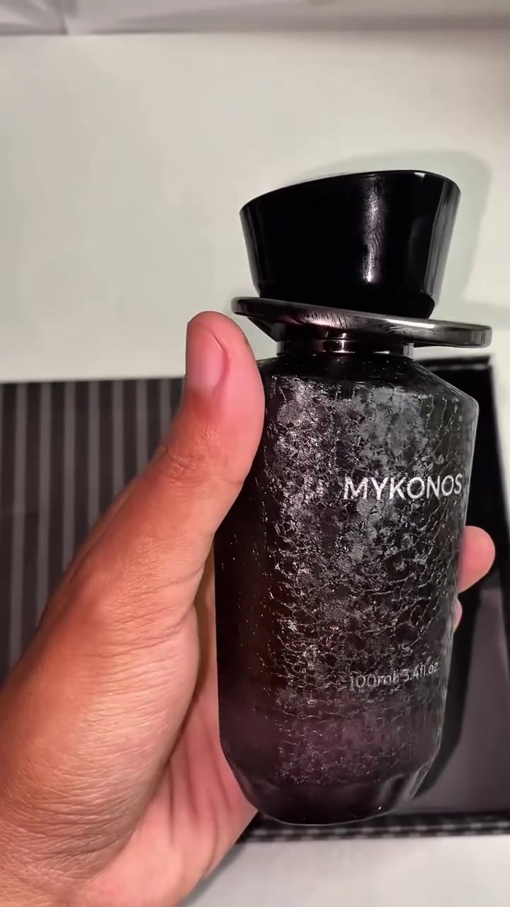 Tangan memegang botol Mykonos Conquer hitam bertekstur ukuran 100 ml dengan tutup hitam mengilap, label Mykonos terbaca jelas