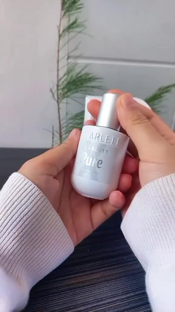 Dua tangan memegang botol Scarlett X Pure berlabel putih di depan ranting tanaman hijau, dengan lengan sweater putih