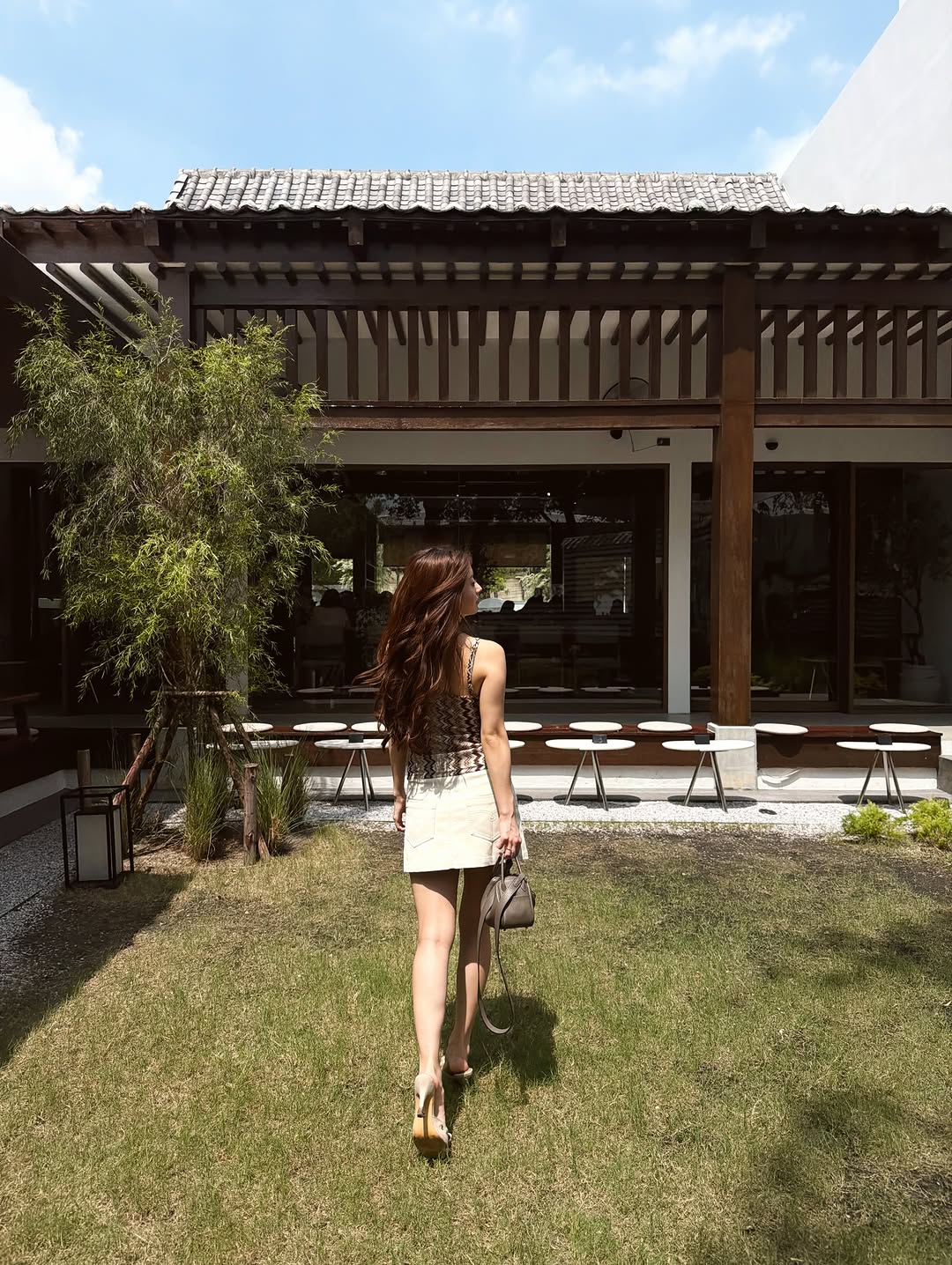 Tampak belakang perempuan berjalan di atas rumput memakai tank top print zigzag, rok mini krem, heels, dan tas jinjing taupe