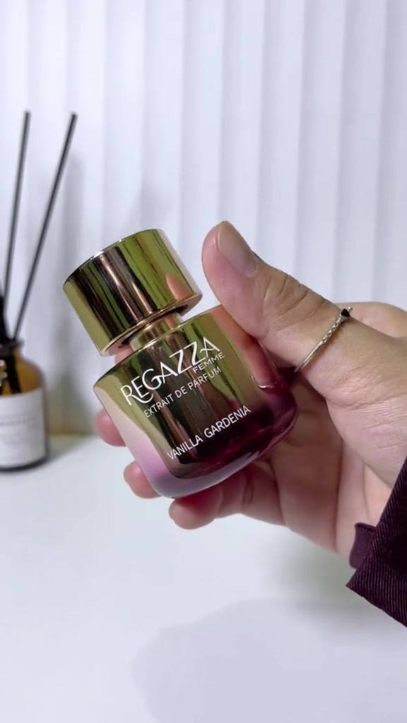 Tangan memegang botol Regazza Femme Extrait de Parfum Vanilla Gardenia bergradasi emas-pink di meja putih dengan diffuser rattan di latar belakang