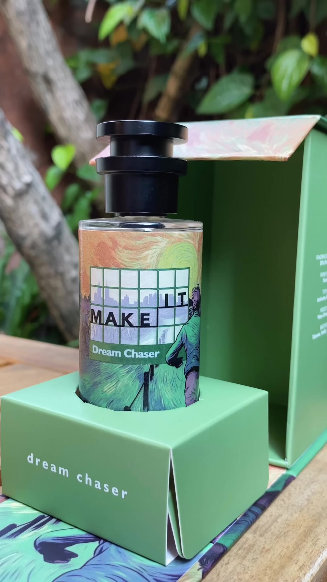Botol Make It Dream Chaser berlabel hijau berdiri di atas kotak hijau di meja kayu luar ruangan dengan dedaunan di belakang