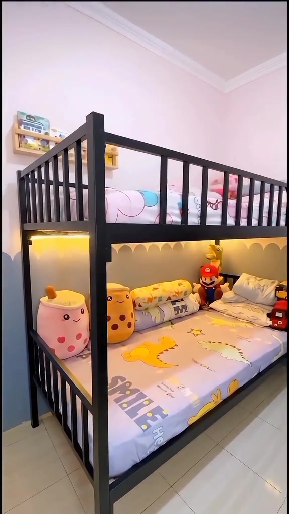 Ranjang tingkat besi hitam di kamar anak berdinding pink dengan seprai motif dinosaurus, bantal guling karakter, dan lampu strip hangat di bawah ranjang atas