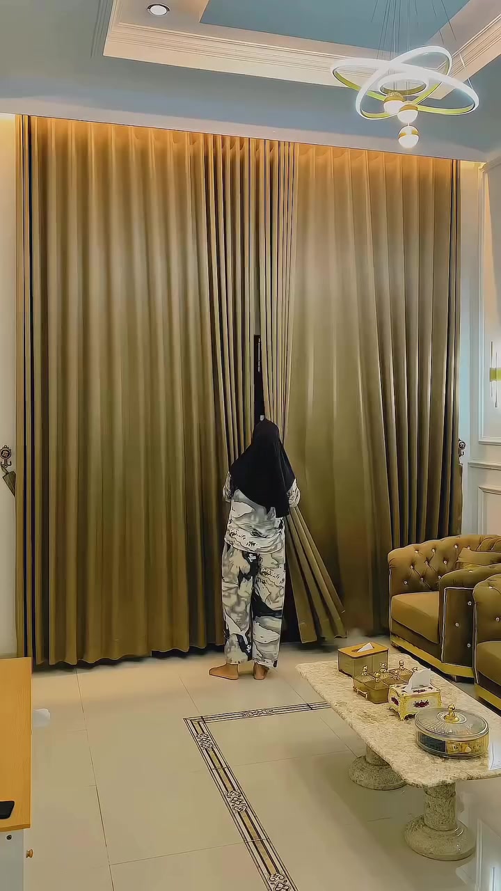 Gorden satin warna emas jatuh dari plafon sampai lantai menutup seluruh dinding jendela sambil dirapikan seseorang dari belakang, dengan meja kopi marmer berisi kotak emas di depannya
