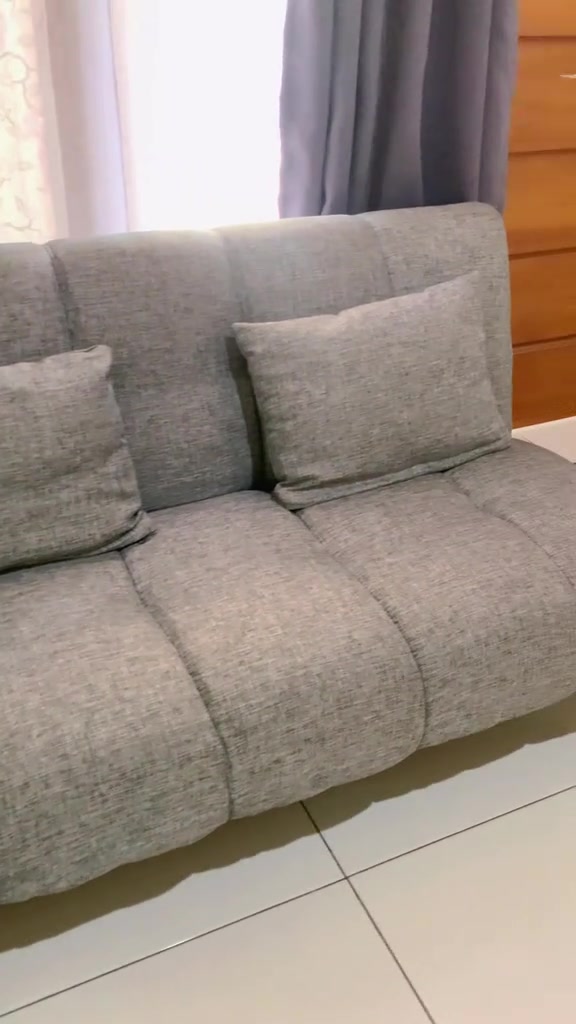Tampak dekat sofa bed kain tenun abu-abu dengan dua bantal sandar di depan gorden dan dinding kayu