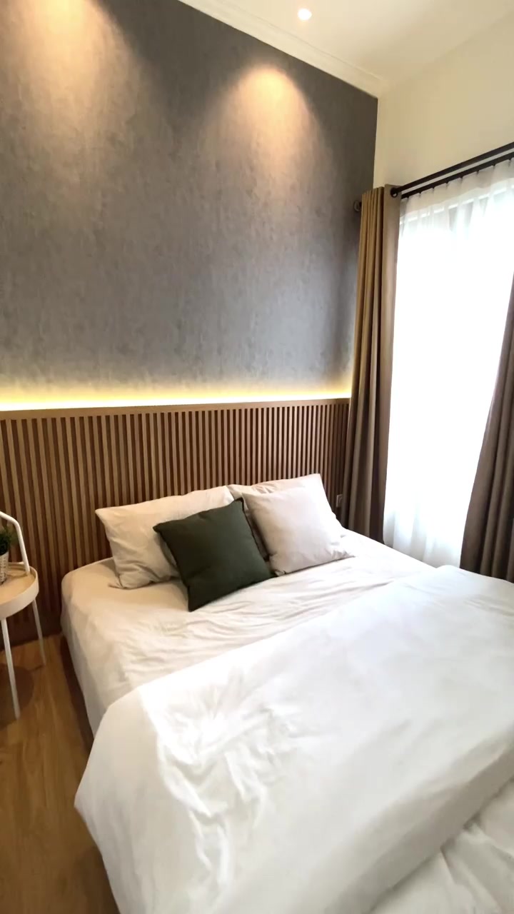 Kamar tidur nyata berdinding abu abu dengan panel kayu fluted di kepala tempat tidur, sprei putih, bantal hijau, dan tirai cokelat di sisi jendela