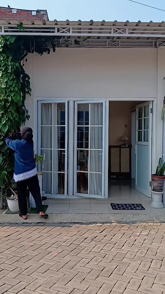Seseorang tampak dari belakang merapikan tanaman rambat di sisi teras rumah berpintu lipat kaca dengan paving blok di depan