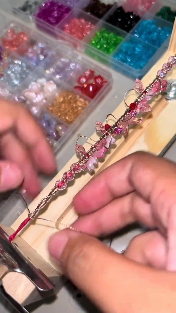 Gelang manik merah muda sedang dirangkai di dudukan kayu dengan kotak manik warna-warni di belakangnya