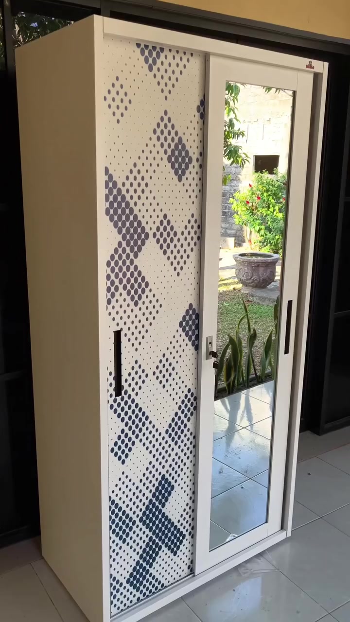 Lemari pakaian pintu geser putih dengan satu pintu bermotif dan satu pintu cermin penuh yang memantulkan taman, berdiri di teras berlantai keramik