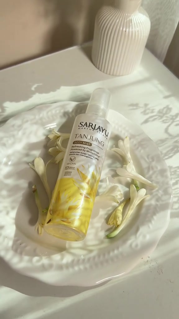 Botol Sariayu Tanjung Body Mist terbaring di piring putih berisi bunga sedap malam dengan cahaya jendela