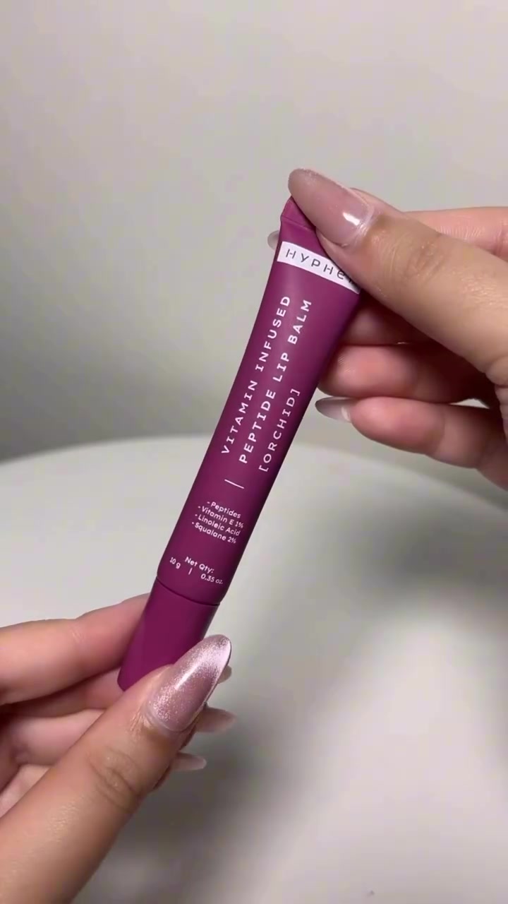 Tangan memegang tube lip balm warna magenta dengan daftar kandungan tercetak di kemasan