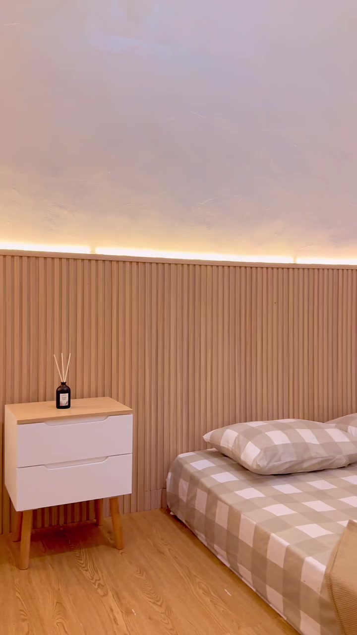 Garis lampu LED hangat menyala di sepanjang ujung atas panel dinding fluted, di samping nakas putih dua laci dan tempat tidur bersprei kotak kotak