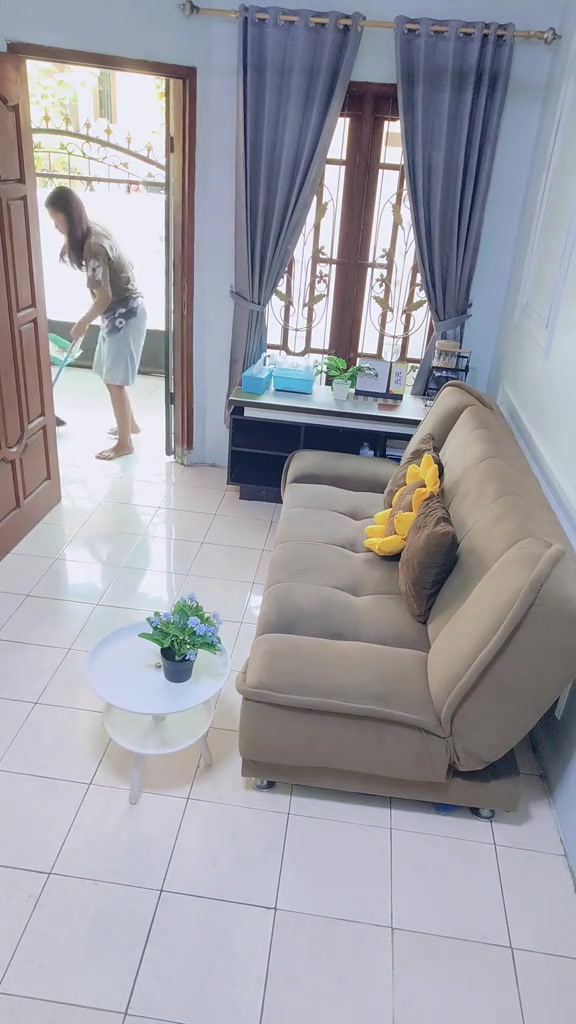 Ruang tamu sempit berlantai keramik dengan sofa bed cokelat muda menempel di dinding, gorden abu-abu, dan meja samping bundar putih