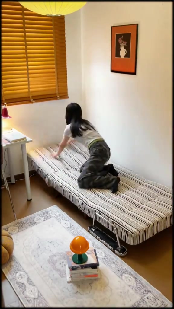 Sofa bed futon motif garis dibentangkan rata menjadi tempat tidur di kamar kecil dengan blind kayu dan karpet motif