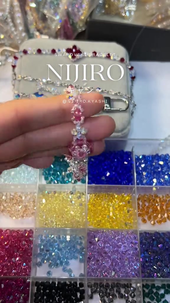 Tangan memegang gelang kristal merah muda motif bunga di atas kotak berisi manik kristal berbagai warna
