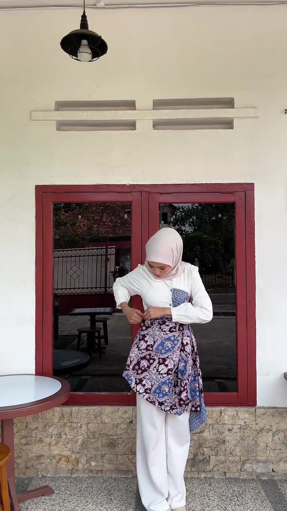 Perempuan berhijab merapikan lilitan kain batik ungu sebagai rok di atas celana putih