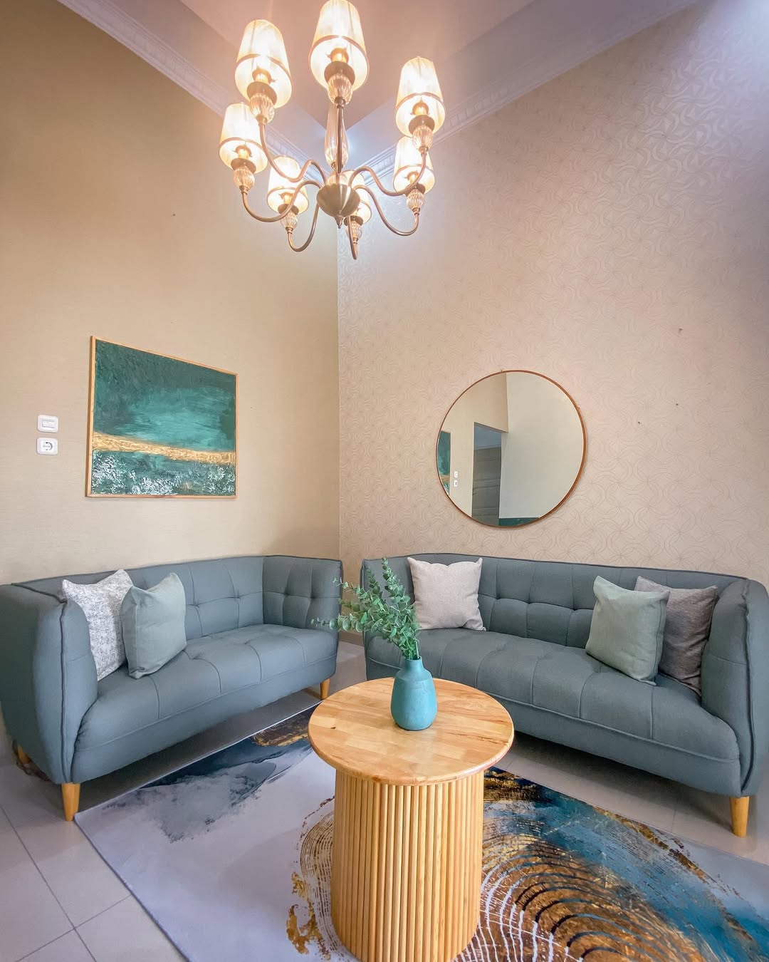 Ruang tamu berdinding wallpaper motif halus warna krem dengan dua sofa hijau sage, meja kayu bundar, dan cermin bulat