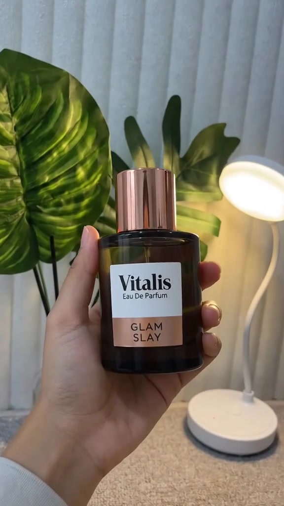 Tangan memegang botol Vitalis EDP Glam Slay berwarna cokelat gelap di depan daun monstera dan lampu meja menyala