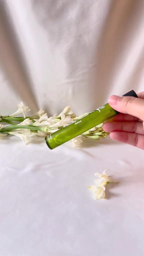 Tangan memegang botol parfum kaca hijau ramping di atas tangkai bunga sedap malam putih