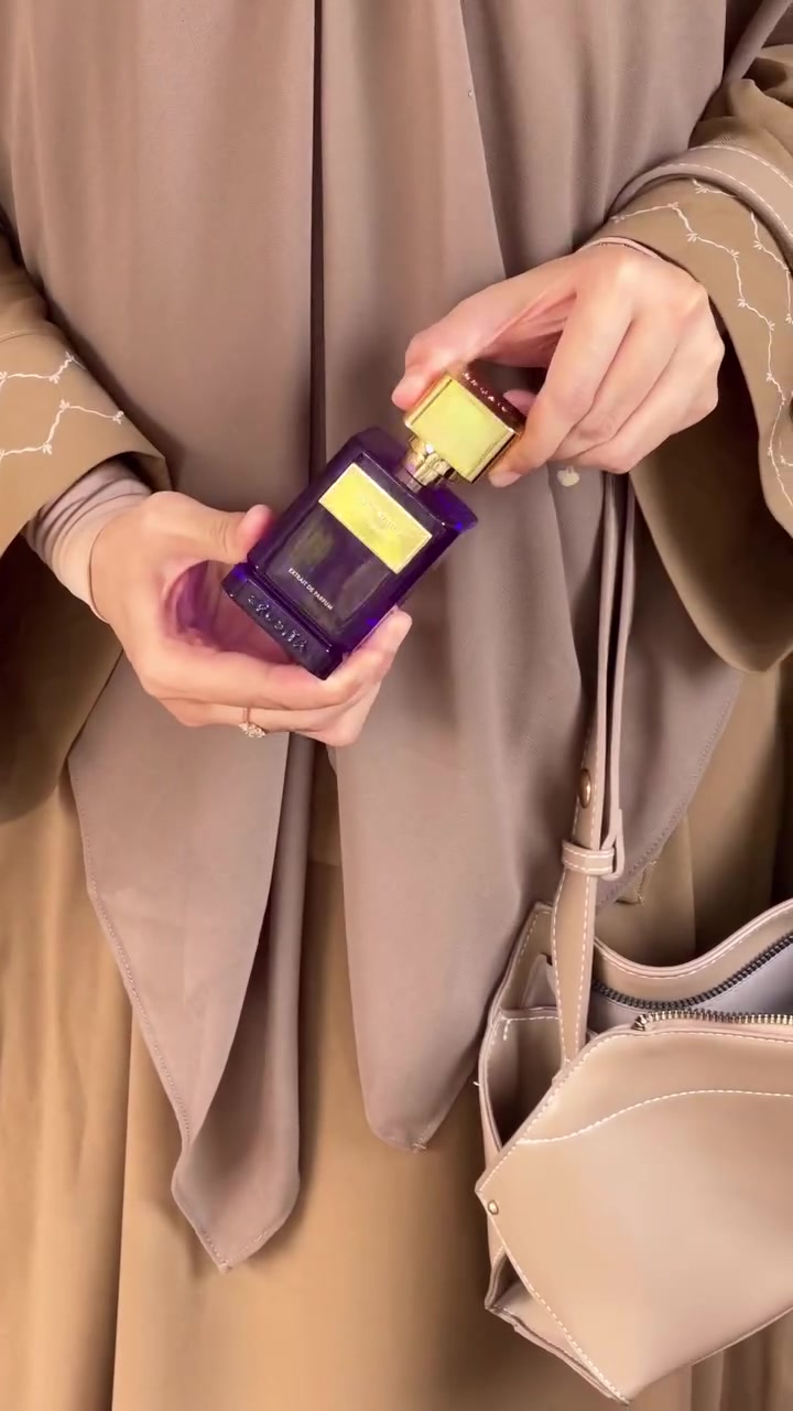 Dua tangan membuka botol parfum ungu Crusita Stuck With U bertutup emas di depan pakaian krem