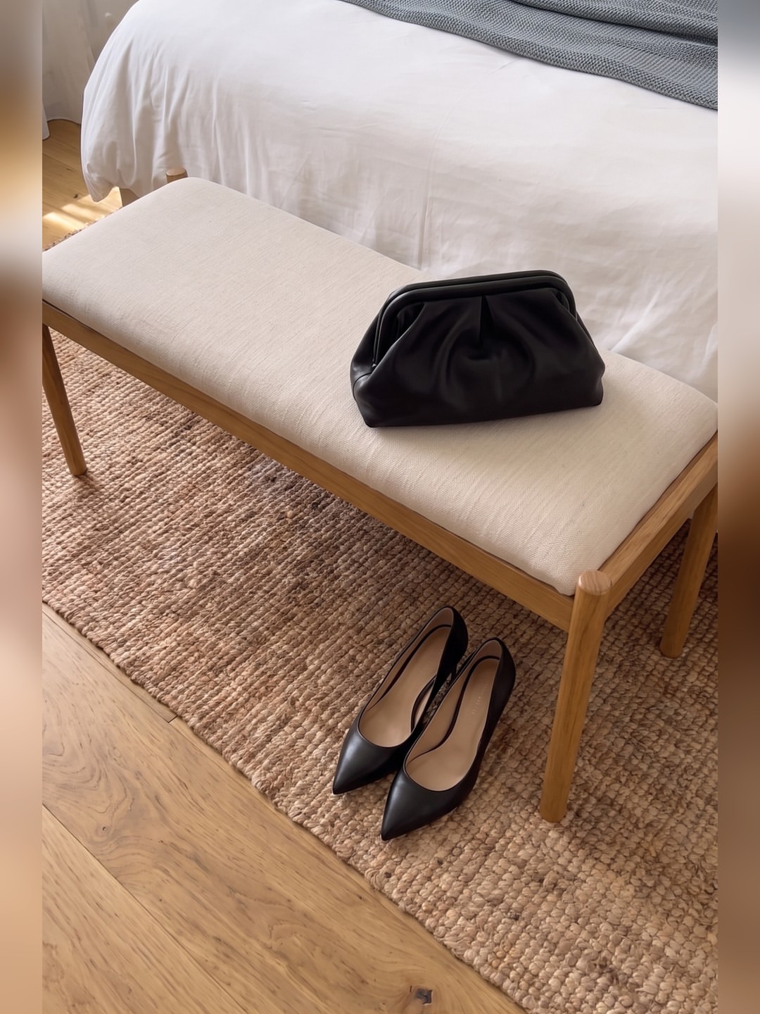 Clutch kulit hitam dan sepasang pumps hitam runcing di atas karut anyaman, di samping bench krem
