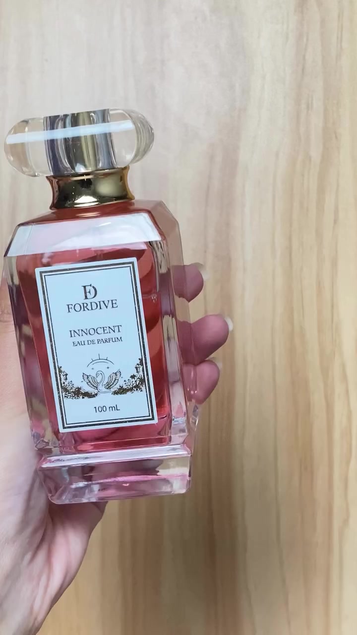 Tangan memegang botol Fordive Innocent eau de parfum 100 mL berisi cairan merah muda di depan latar kayu terang