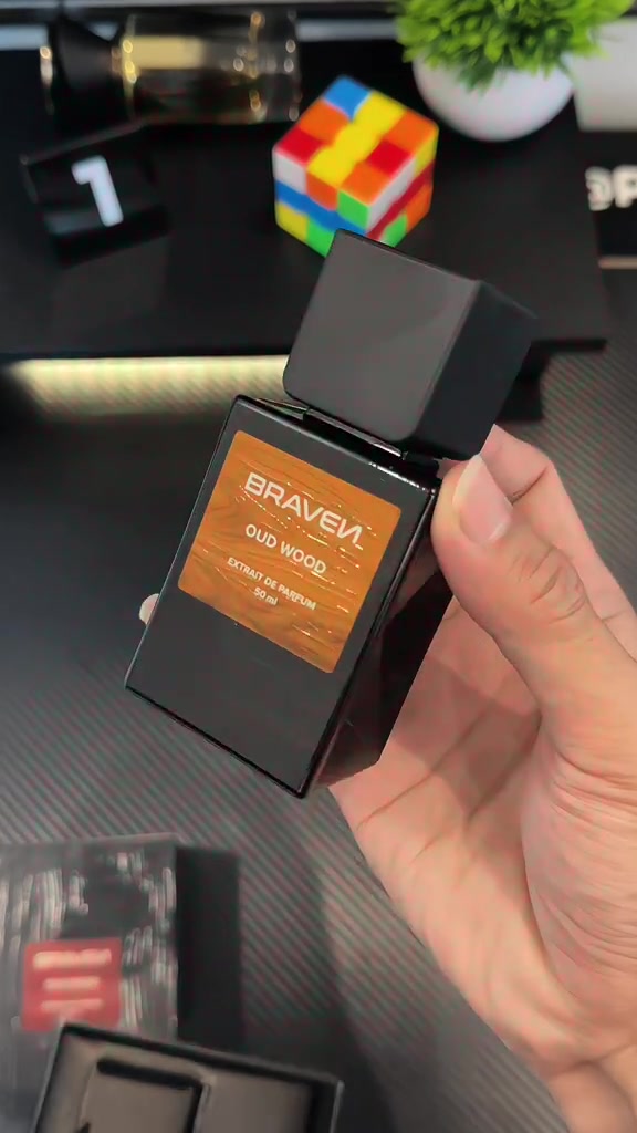 Tangan memegang botol Braven Oud Wood extrait 50 ml dengan kotak hitam, tanaman, dan rubik di latar belakang