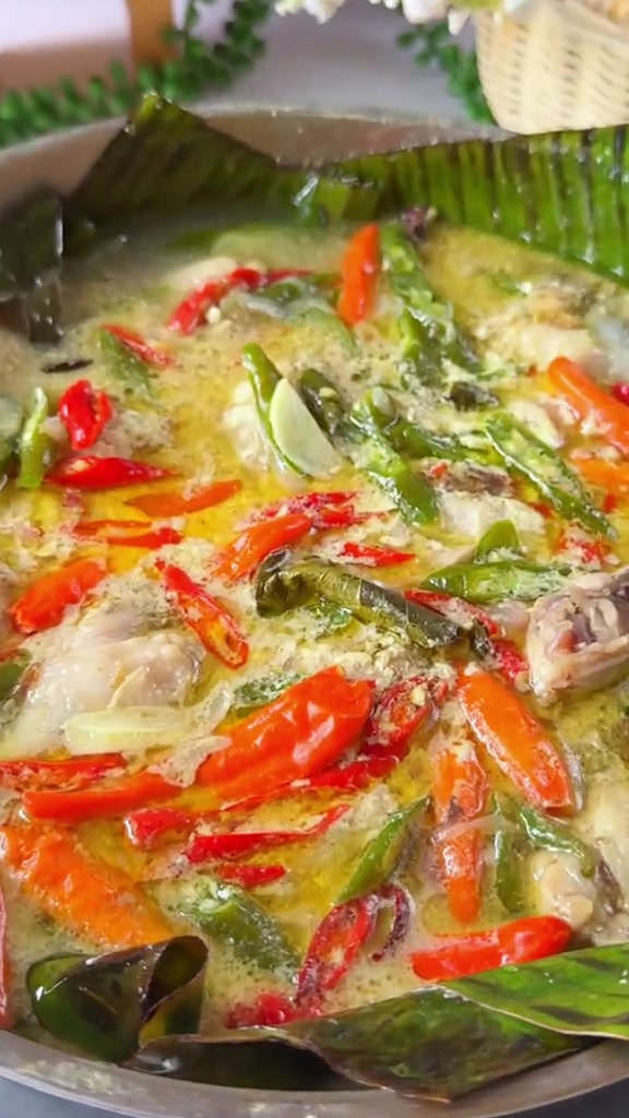 Panci lebar berisi garang asem ayam berkuah santan mengilap dengan cabai merah, cabai hijau, dan lembaran daun pisang di tepi panci