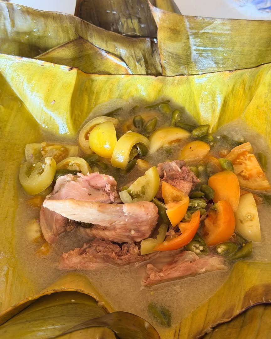 Garang asem ayam disajikan di atas daun pisang dengan potongan tomat hijau, tomat oranye, dan cabai iris, kuahnya terlihat segar