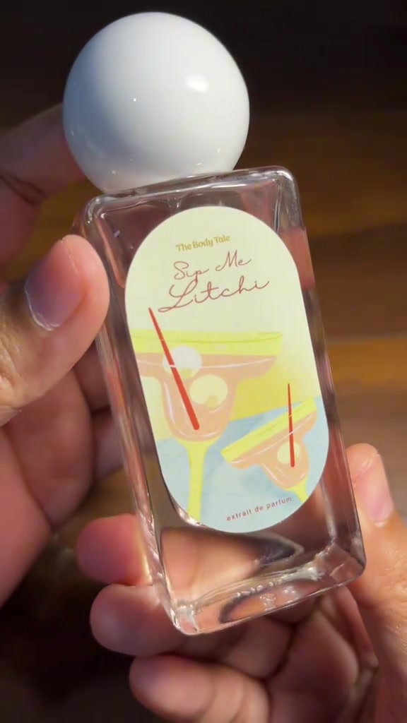 Tangan memegang botol The Body Tale Sip Me Litchi berlabel putih di depan latar kayu hangat