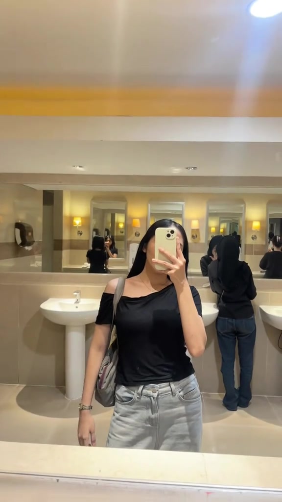 Perempuan memakai kaos one shoulder hitam dan jeans abu-abu muda memotret diri di cermin toilet umum, wajah tertutup ponsel