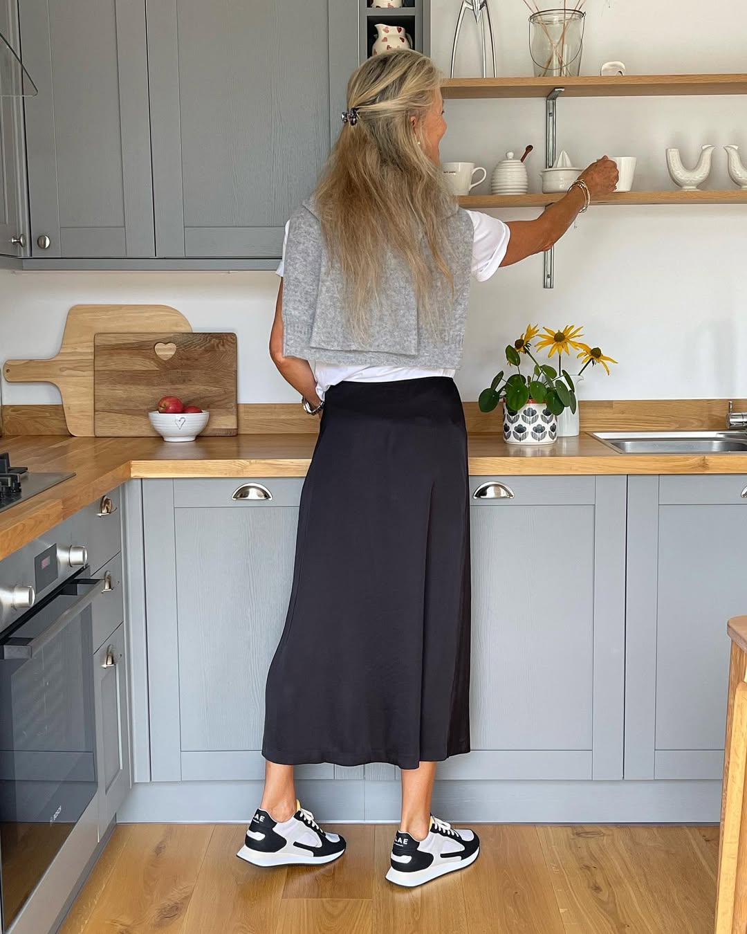 Tampak belakang wanita di dapur memakai slip skirt satin hitam panjang, kaos putih, sweater abu-abu disampir di bahu, dan sneakers putih hitam