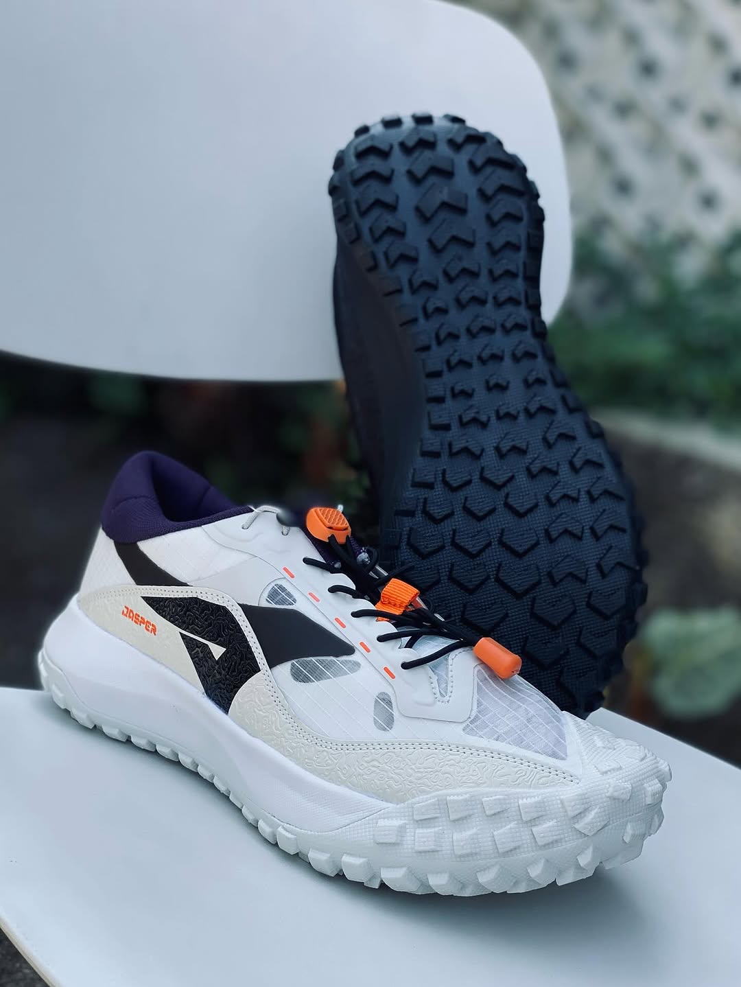 Outsole Ardiles ATF Jasper menghadap kamera memperlihatkan pola lug dua arah, di depannya sepatu colorway putih