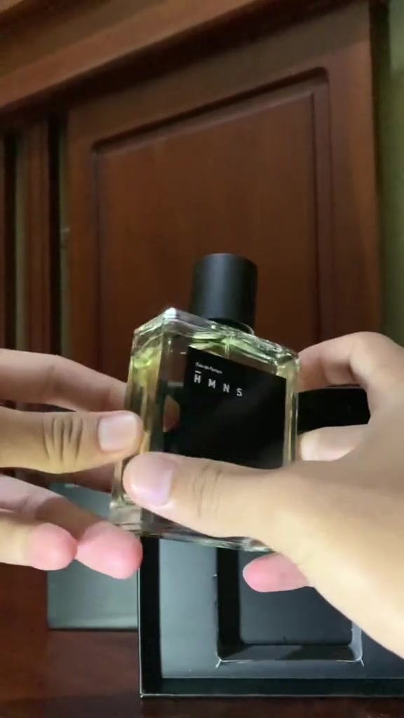 Dua tangan memegang botol HMNS Alpha berisi cairan hijau muda di depan kotak hitamnya