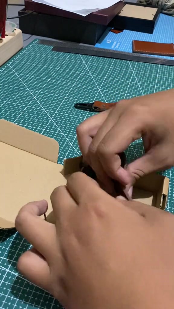 Tangan pengrajin merapikan keychain kulit di atas talenan potong hijau dengan strap kulit dan kait lobster terlihat di meja