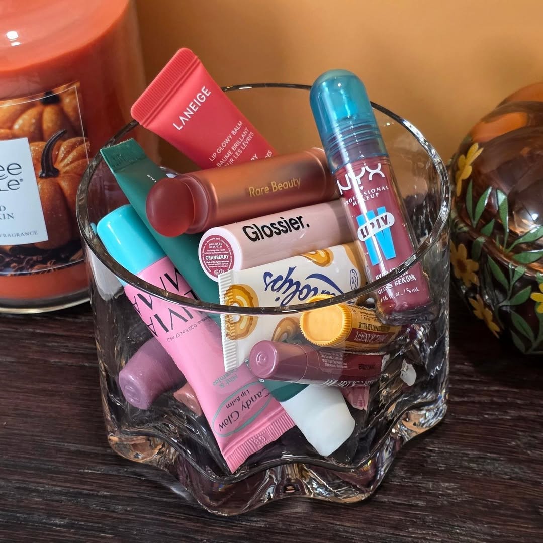 Berbagai lip balm dan lip oil, termasuk NYX lip oil, terkumpul dalam wadah kaca di atas meja kayu