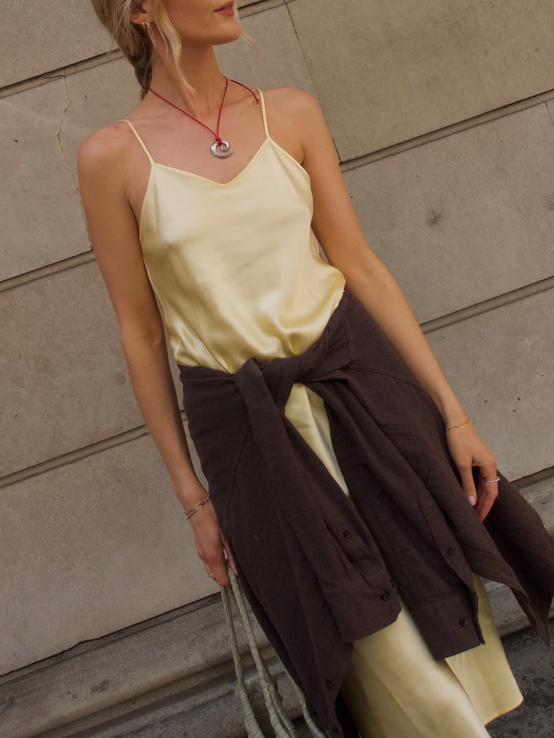 Detail dekat slip dress satin kuning mentega berkerah cowl dengan kemeja cokelat diikat di pinggang dan tas rajut