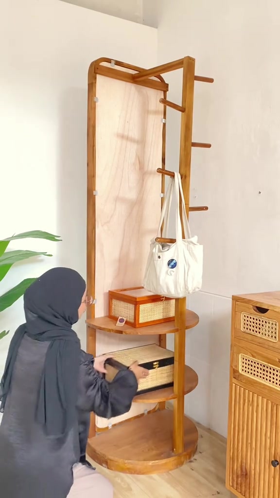 Sisi belakang cermin putar berbingkai kayu dengan rak bulat bertingkat dan tiang gantungan tempat tas kain tergantung, seseorang berjongkok menata kotak di rak bawah