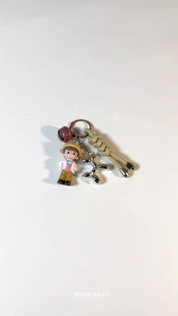 Bag charm berisi figure anak kecil bertopi, charm logam balon anjing, lonceng cokelat, dan simpul paracord hijau di atas latar putih