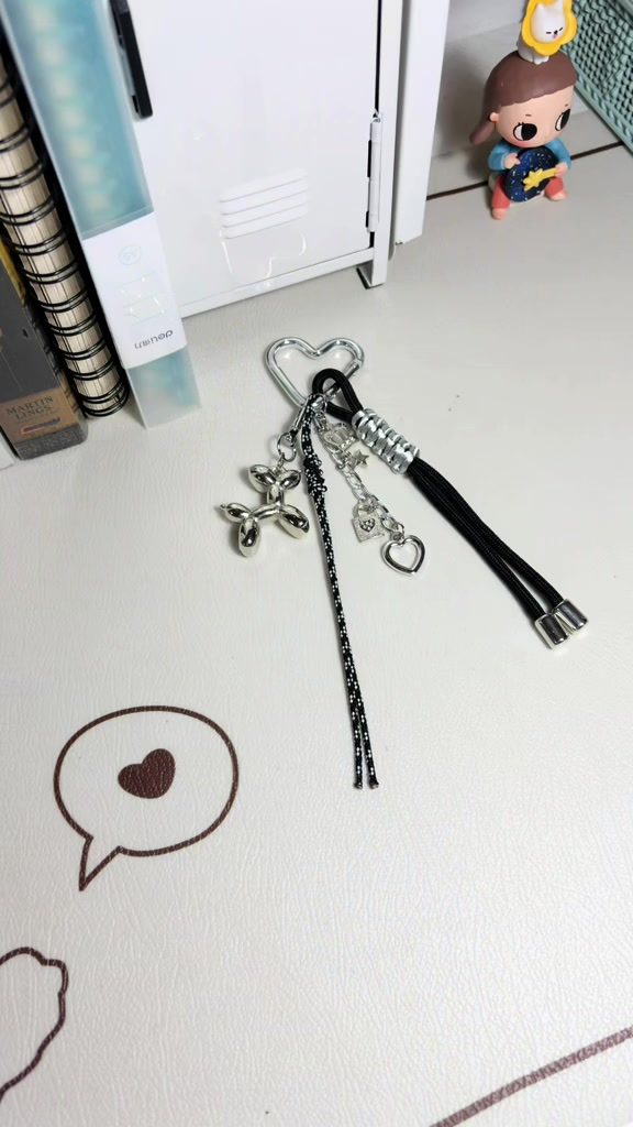 Bag charm dengan kait karabiner bentuk hati, charm logam balon anjing, rantai bintang, dan tali hitam tergeletak di atas meja putih