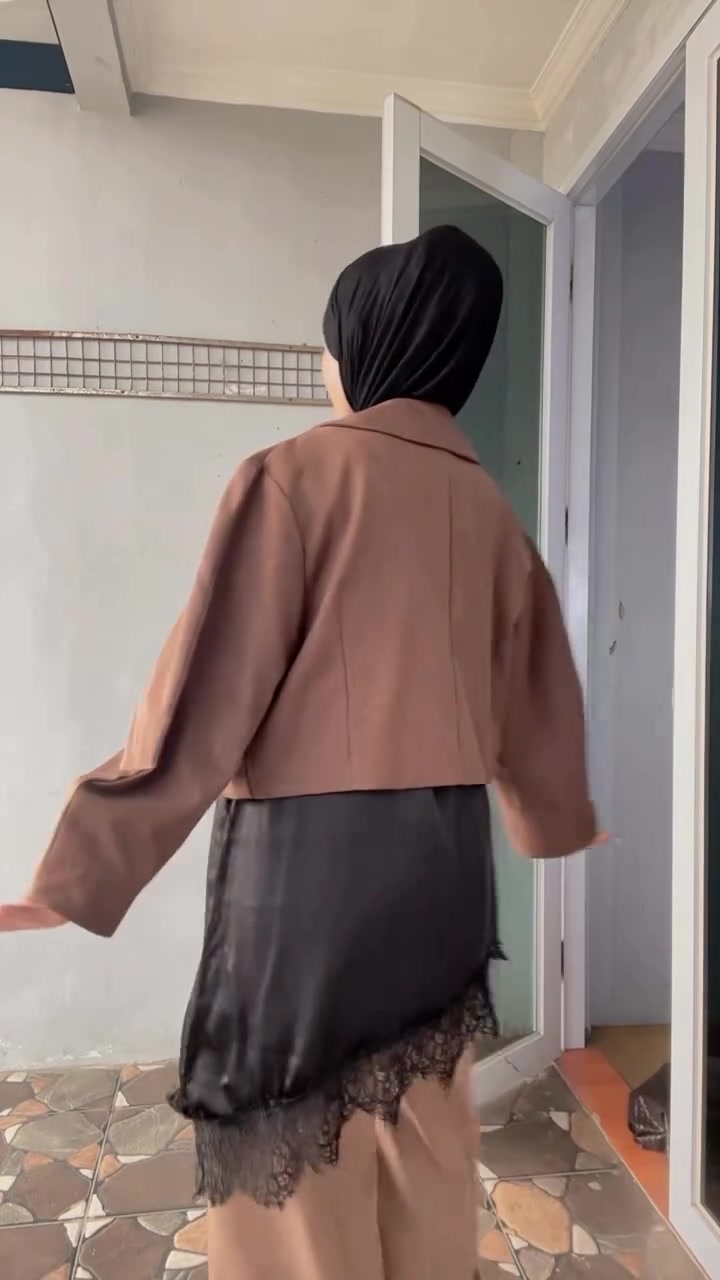 Tampak belakang perempuan berhijab hitam memakai outer cropped cokelat berlengan balon di atas inner lace satin hitam dan celana longgar beige