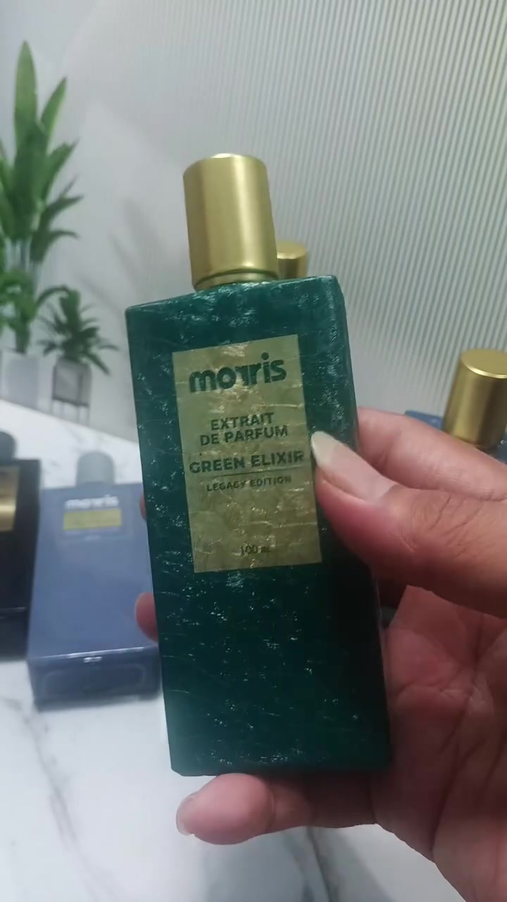 Botol Morris Legacy Edition Green Elixir berwarna hijau teal dipegang di atas meja marmer dengan tanaman dan botol Morris lain di latar belakang