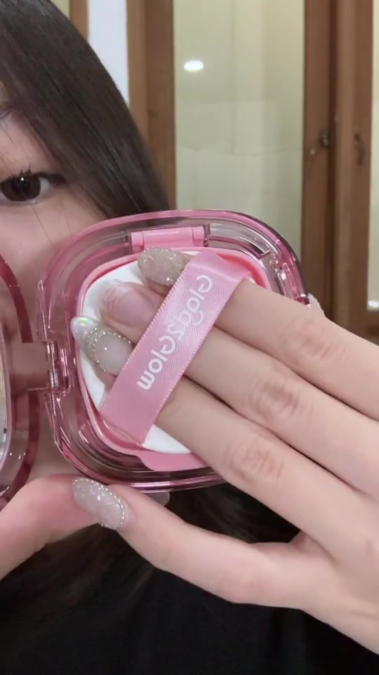 Tangan dengan puff bertali pink di atas kompak cushion transparan berisi produk, posisi siap menempelkan produk dengan cara tap