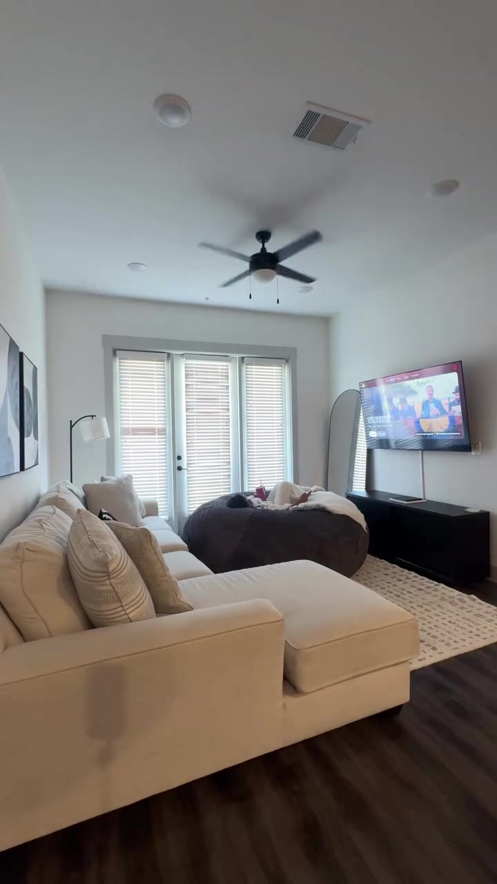 Ruang keluarga terang dengan sofa sectional krem dan beanbag jumbo cokelat tempat seseorang berbaring di dekat jendela