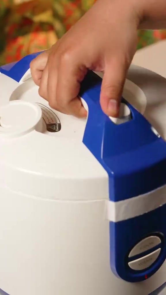 Tangan menekan tombol cook pada rice cooker putih biru saat memasak nasi daun jeruk
