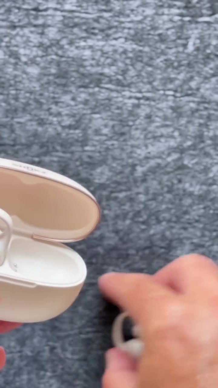Casing charging warna krem terbuka dengan earbuds open-ear clip-on sedang diambil dengan tangan