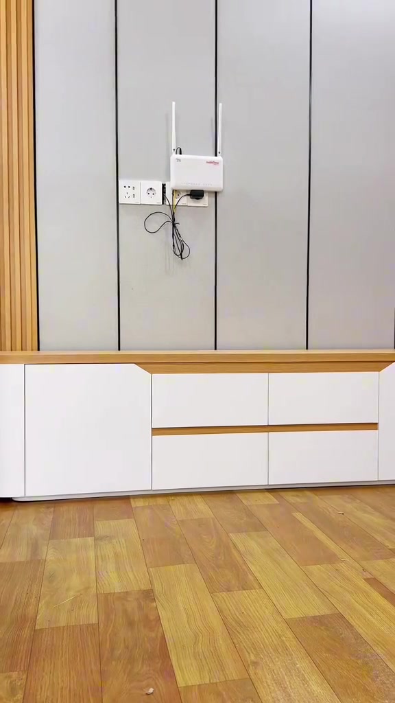 Meja TV minimalis putih dengan top kayu di bawah dinding panel abu-abu, dengan router dan kabel yang masih terjuntai di dinding