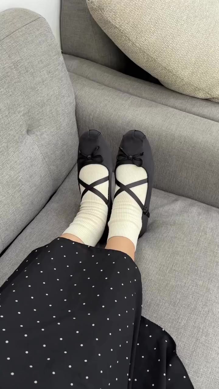 Mary jane satin hitam berstrap silang dipakai dengan kaos kaki cream dan rok polkadot, kaki selonjoran di sofa abu-abu