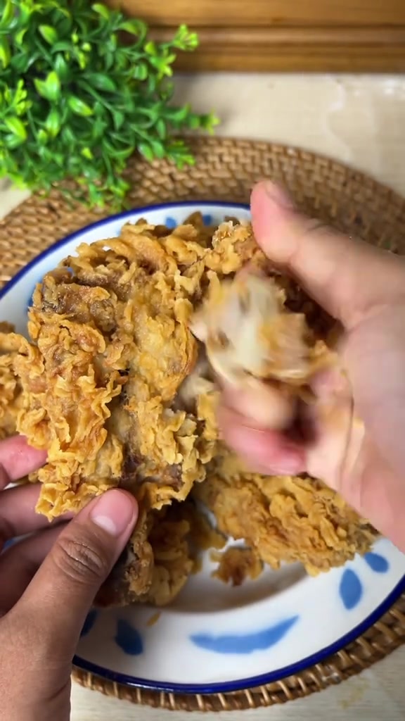Tumpukan ayam goreng tepung keriting keemasan di piring bermotif biru, satu potongan disobek memperlihatkan daging putih yang juicy