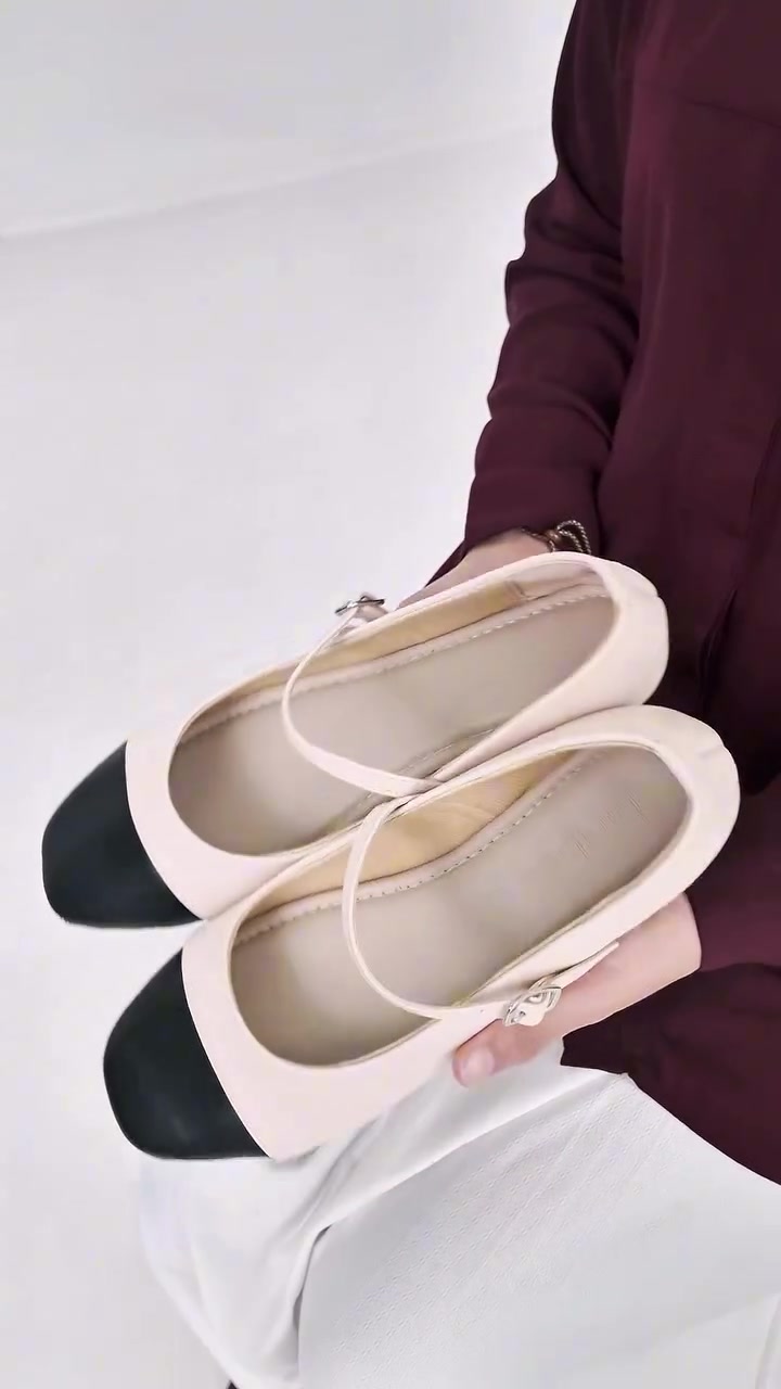 Tangan memegang sepasang mary jane cap-toe warna cream dengan ujung hitam dan strap berbuckle