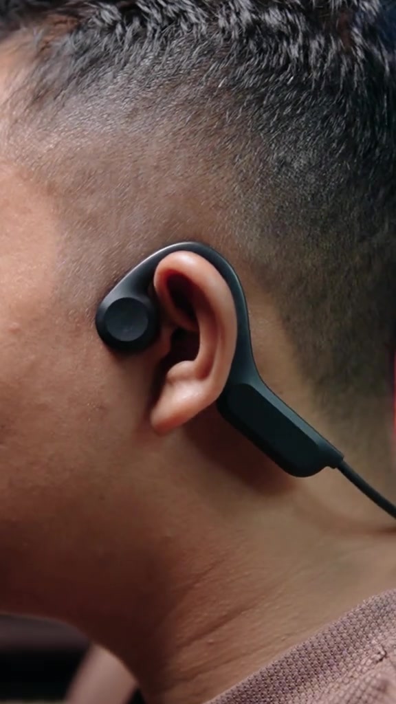 Headset bone conduction hitam tampak samping terpasang mengait di telinga dengan pad di depan lubang telinga
